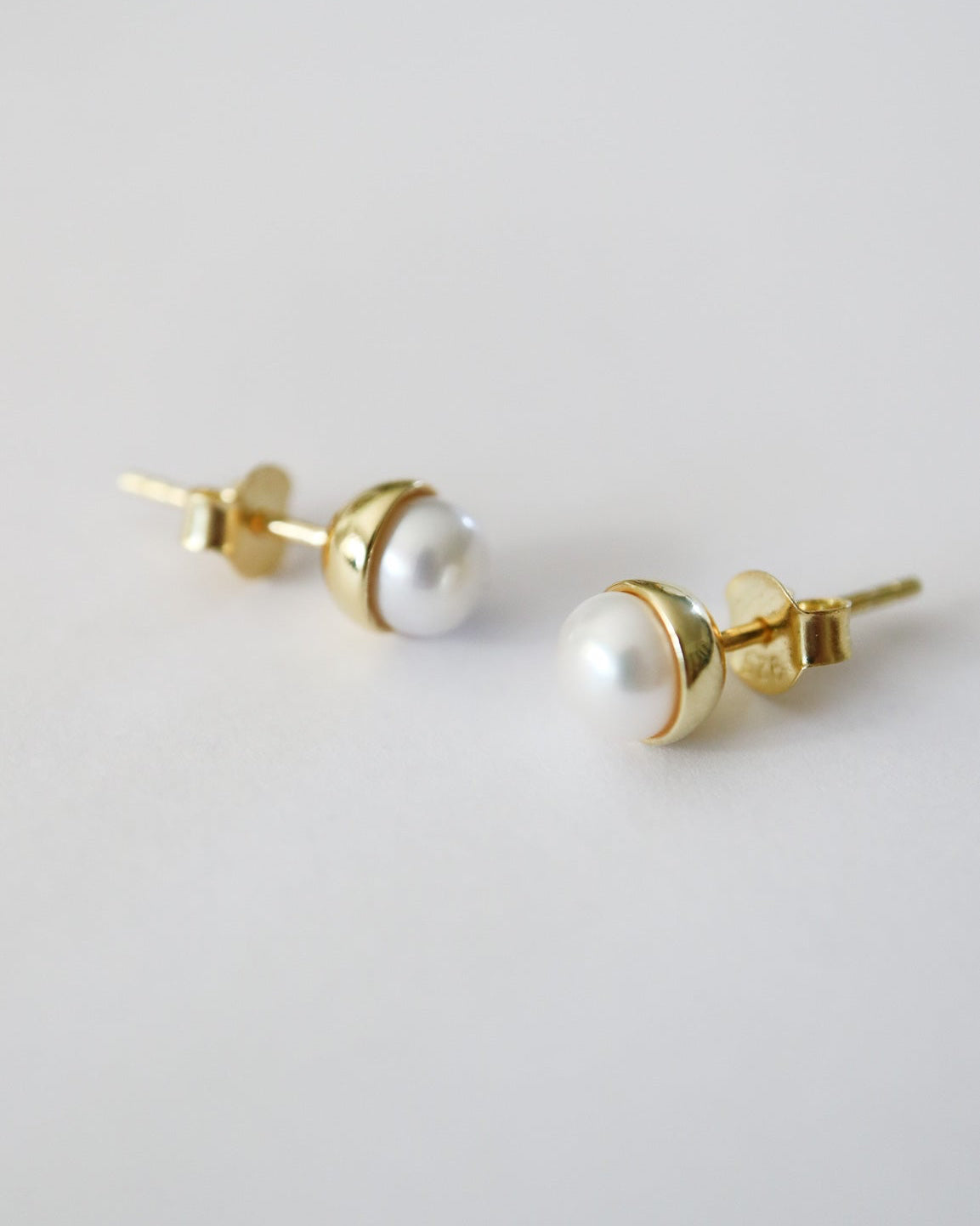 A pair of gold stud earrings with white freshwater pearls, shown from a side angle on a clean, light gray background.