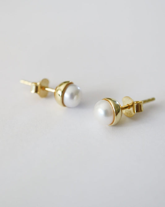 A pair of gold stud earrings with white freshwater pearls, shown from a side angle on a clean, light gray background.
