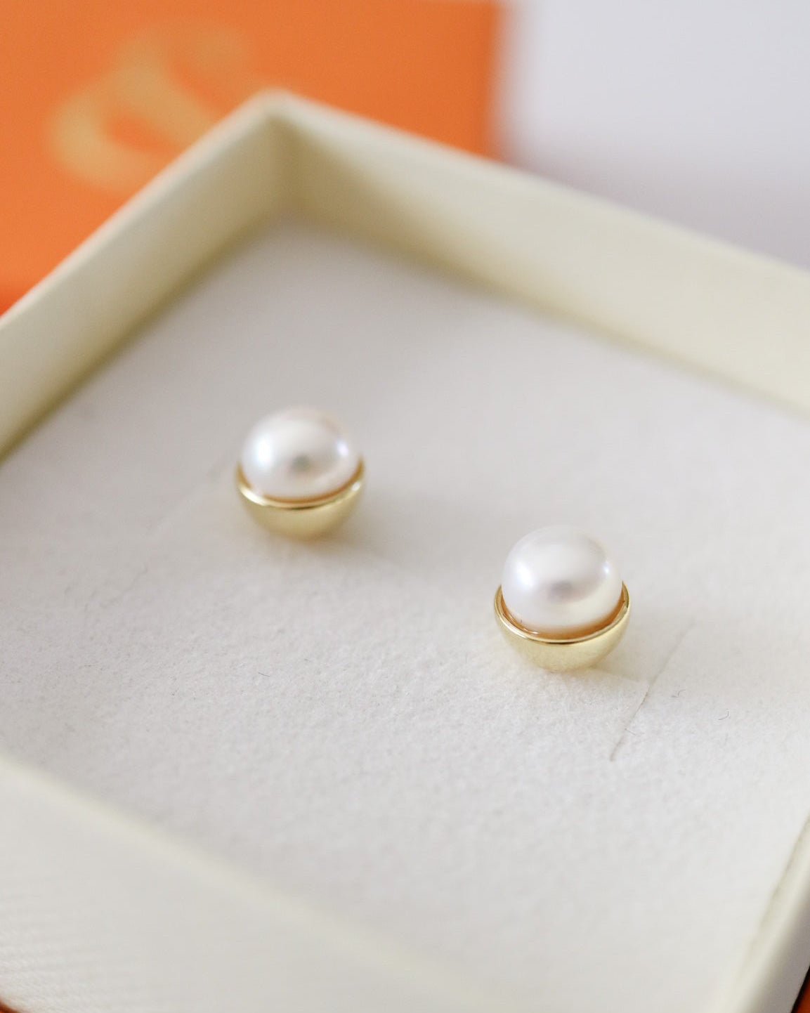 A pair of gold stud earrings with white pearl-like beads, displayed inside an open cream-colored jewelry box with an orange lid in the background.