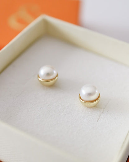 A pair of gold stud earrings with white pearl-like beads, displayed inside an open cream-colored jewelry box with an orange lid in the background.