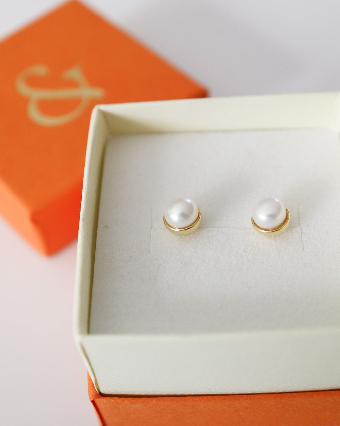 A pair of gold stud earrings with white pearl-like centers, displayed in an open cream jewelry box with an orange lid in the background.