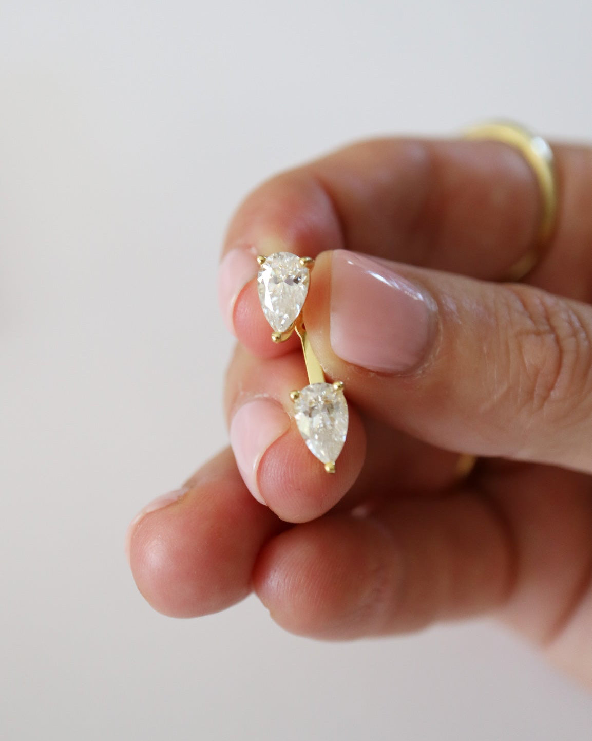A close-up of a hand holding a pair of gold earrings with pear-cut clear gemstones, with a gold band visible on the ring finger against a soft light background.