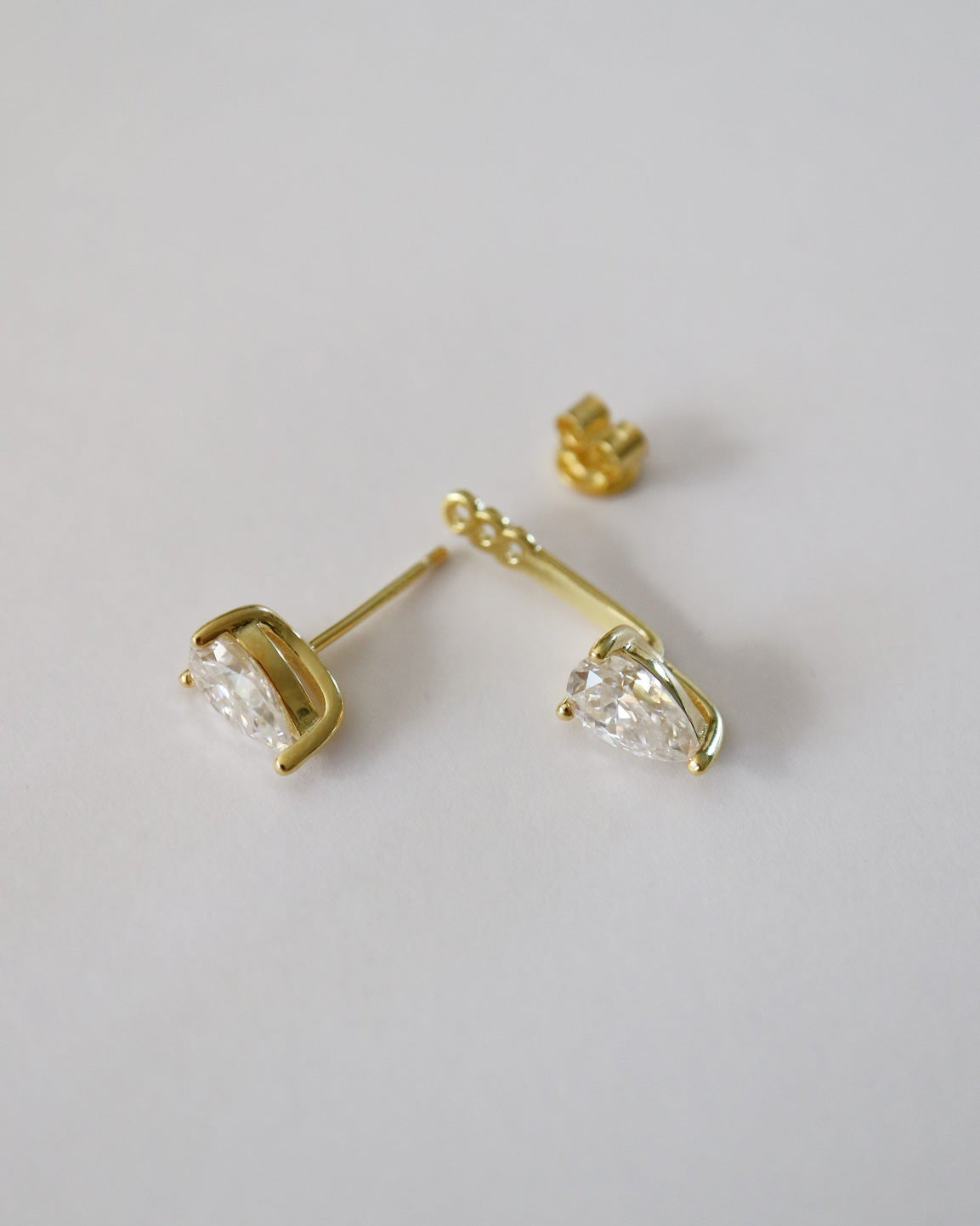 A pair of gold stud earrings with marquise-cut clear gemstones, shown on a plain light gray background. One earring is displayed in profile, showing the post and back, while the other is angled to show the front of the stone and its gold setting.