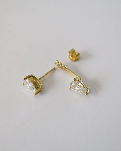A pair of gold stud earrings with marquise-cut clear gemstones, shown on a plain light gray background. One earring is displayed in profile, showing the post and back, while the other is angled to show the front of the stone and its gold setting.