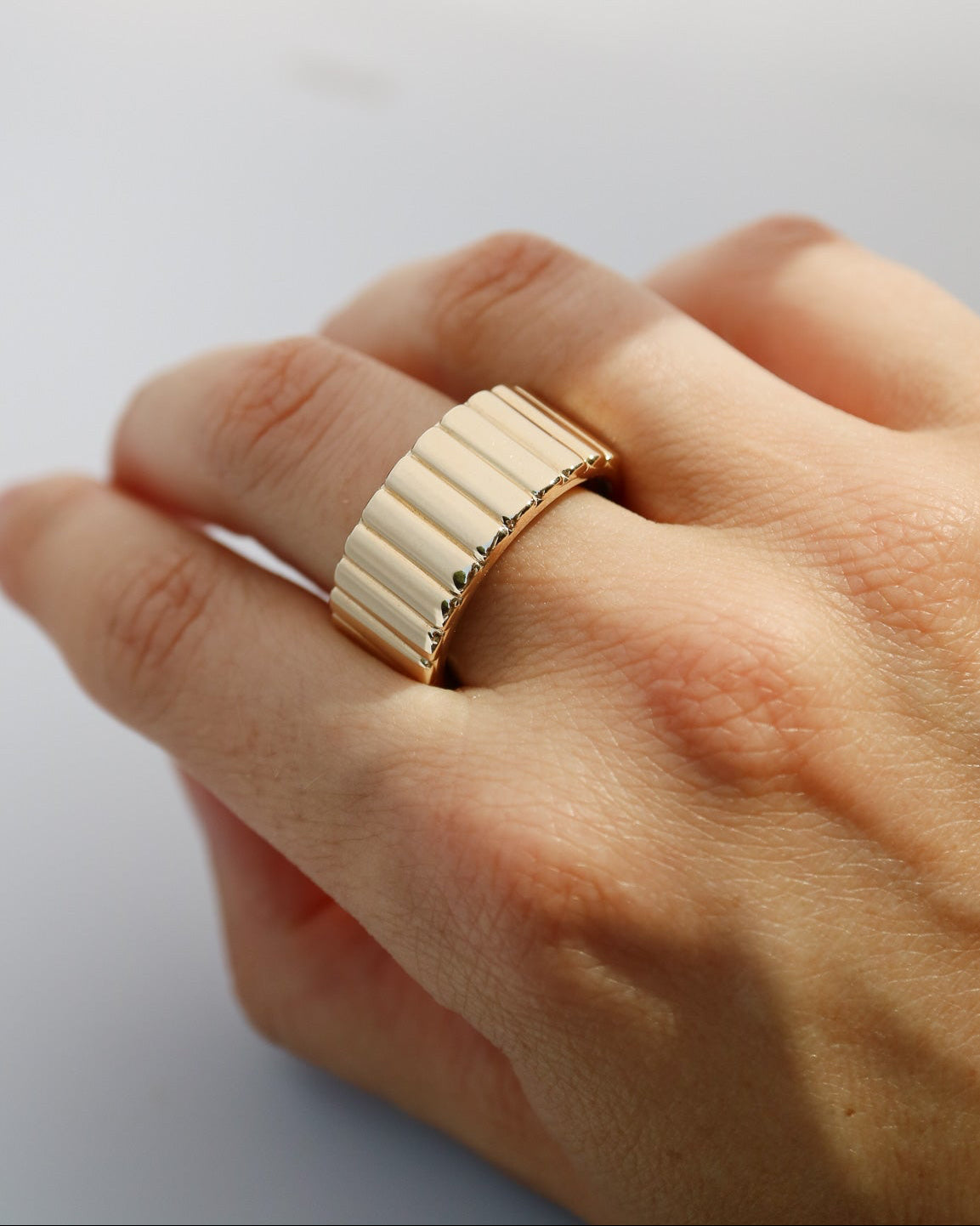 A close-up of a hand wearing a wide, ribbed gold band ring against a light gray background.
