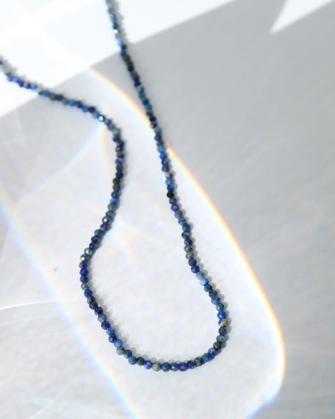 A delicate dark blue beaded chain necklace, likely made of seed beads, laid out on a light gray surface with dramatic light rays and shadows.