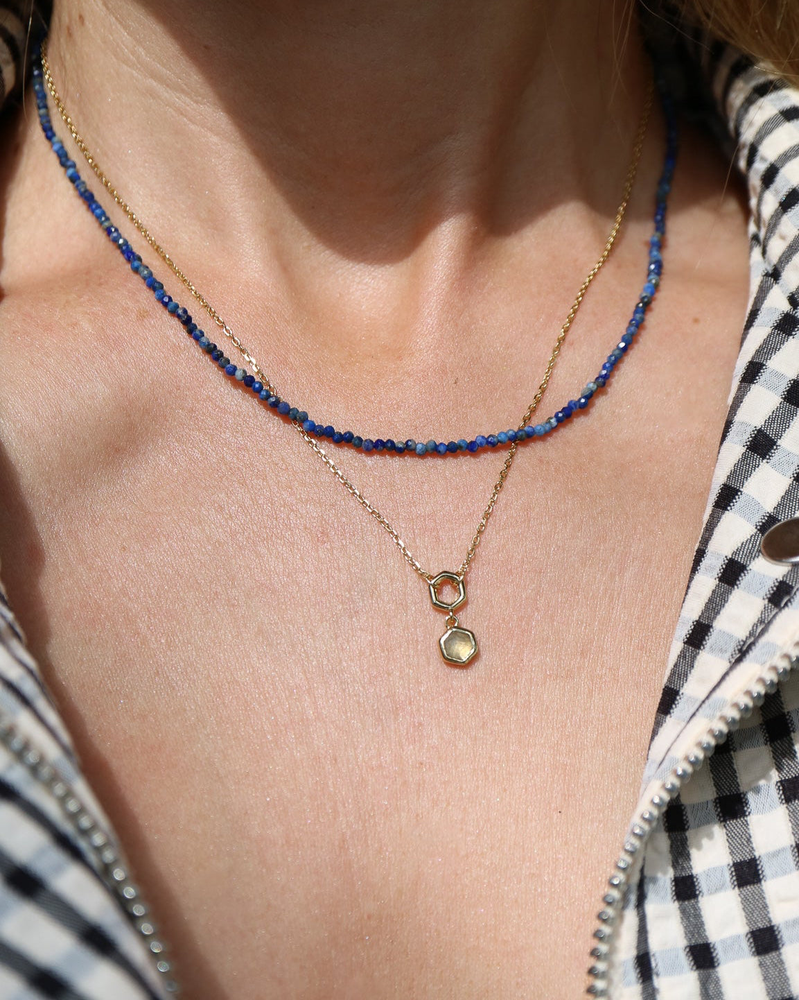 A close-up of a person's neck and chest wearing two layered necklaces: one with small dark blue beads and one gold chain with a double-ring pendant and a round light-colored stone charm. The person is wearing an open black-and-white gingham zip jacket over a white top.