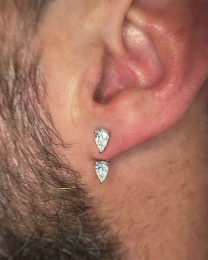 Close-up of a person's ear wearing two small pear-cut clear stud earrings in silver-toned settings.