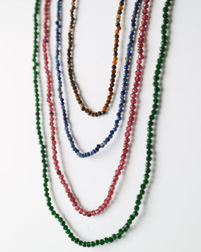 Five beaded strand necklaces in green, red, blue, brown, and orange, draped over a white display form against a white background.