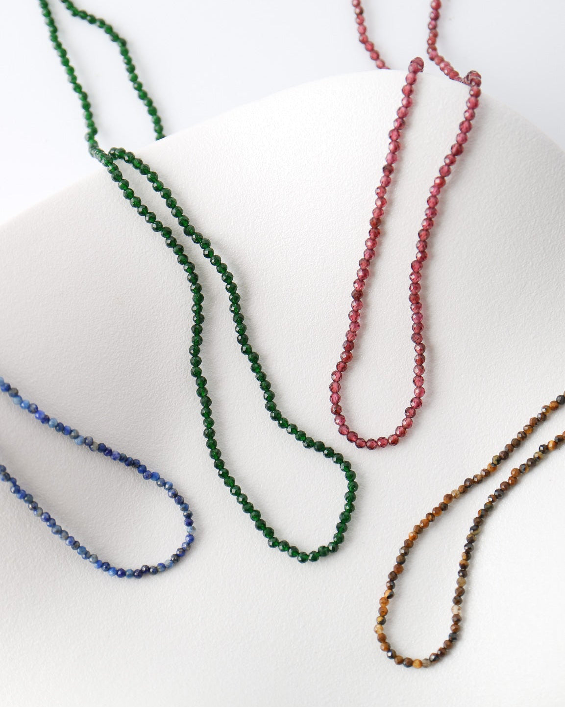 Four beaded chain necklaces in green, red, blue, and brown tones arranged on a white curved display form.