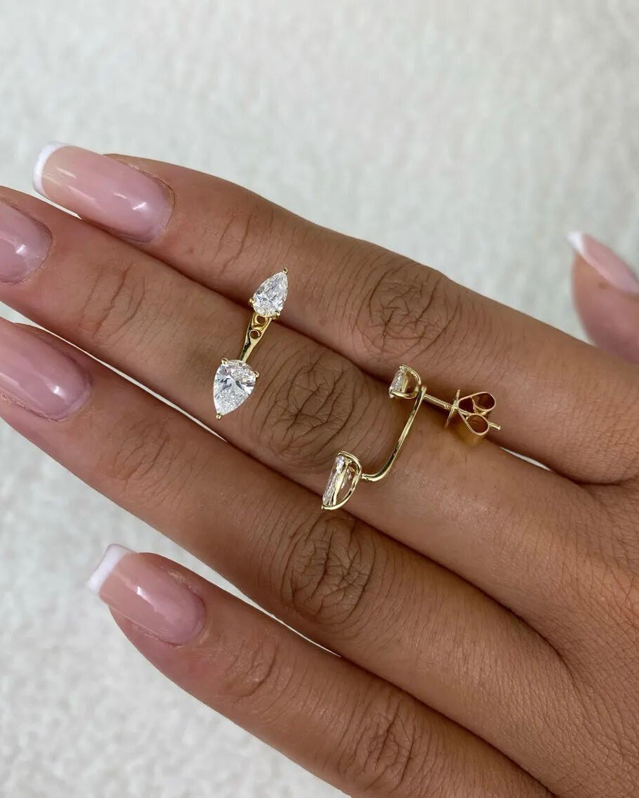 A close-up of a hand with a French manicure wearing two gold huggie hoop earrings, each featuring a dangling clear heart-shaped crystal.