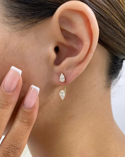A close-up of a person's ear wearing a pair of gold dangle earrings with pear-cut clear stones, with manicured nails resting near the cheek.