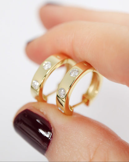 Gold hoop earrings with embedded stones on a hand with dark nail polish.