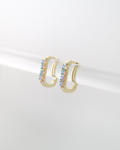A pair of gold huggie hoop earrings with a row of iridescent, rainbow-colored stones on the front, displayed on a white cylindrical stand against a light gray background.