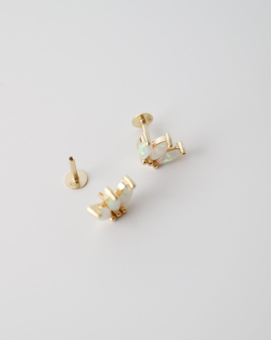 A pair of gold-toned stud earrings with pale iridescent inlay, shown on a clean light gray background. One earring is displayed in front, and the other is turned sideways to show the post and backing.