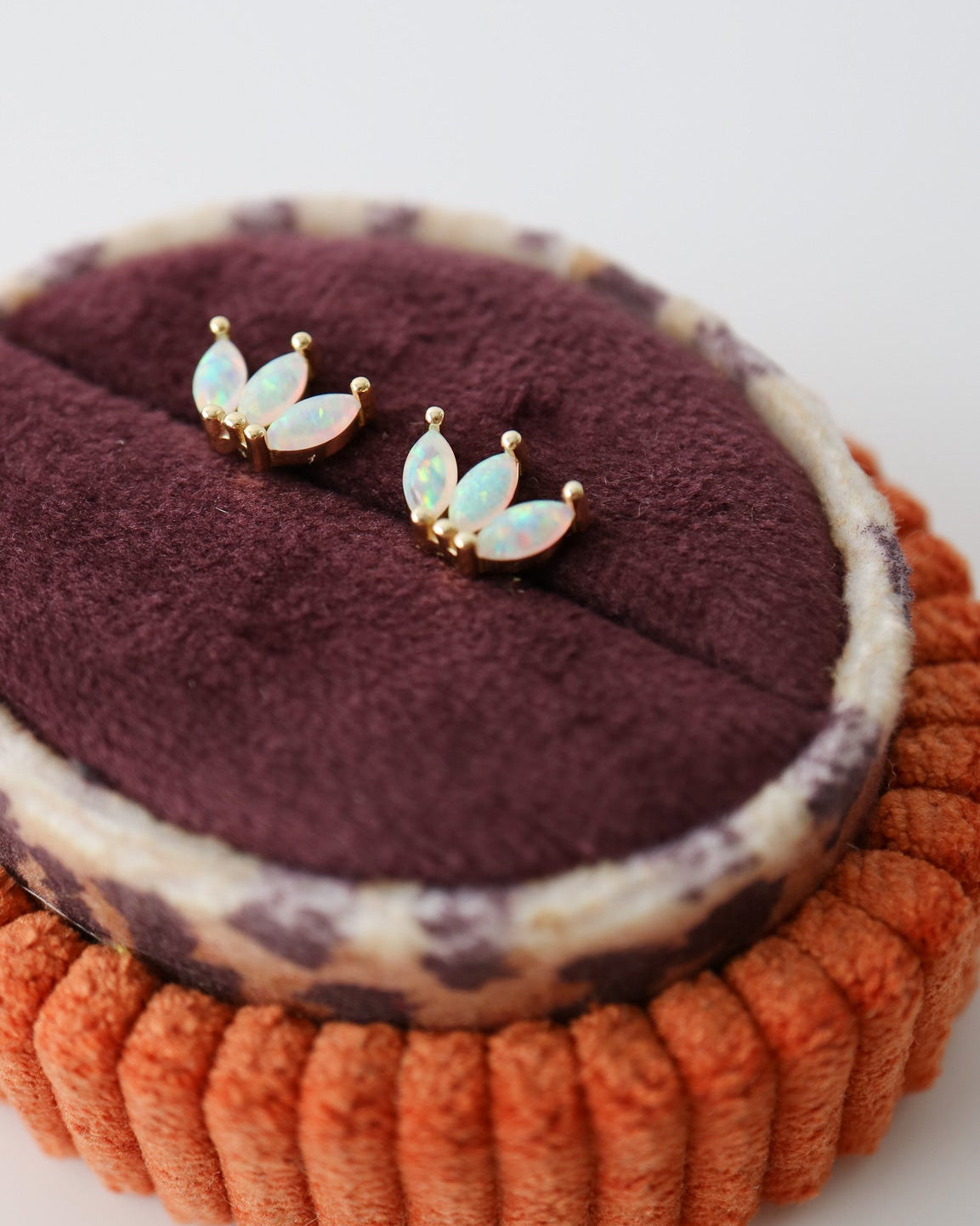 A pair of small gold-tone stud earrings with iridescent white opal inlays, displayed on a dark burgundy velvet jewelry pad atop an orange textured ring stand against a light gray background.