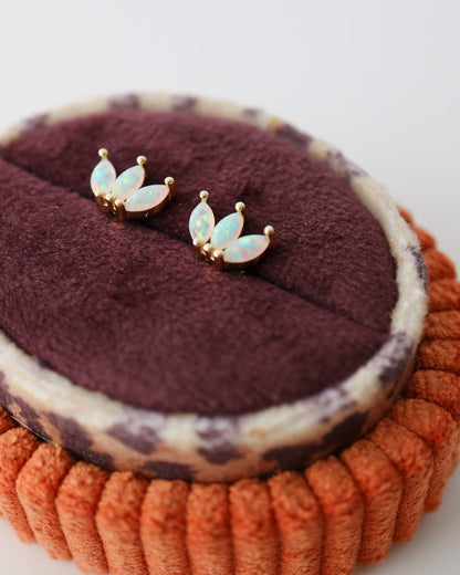 A pair of small gold-tone stud earrings with iridescent white opal inlays, displayed on a dark burgundy velvet jewelry pad atop an orange textured ring stand against a light gray background.