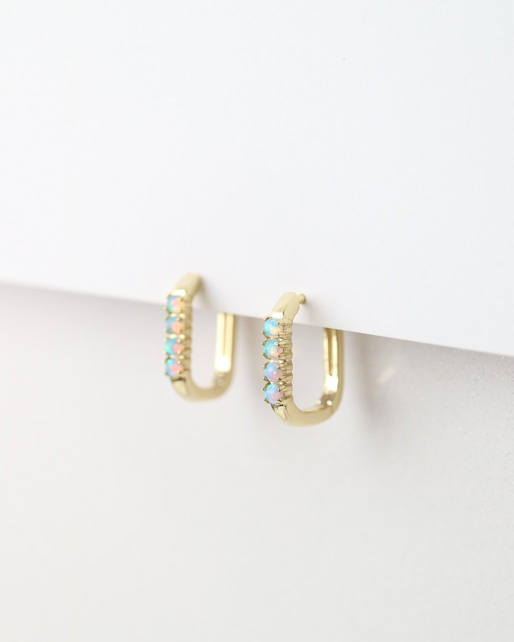 A pair of small gold huggie hoop earrings with rows of iridescent, multicolored opal stones, displayed against a clean white background.