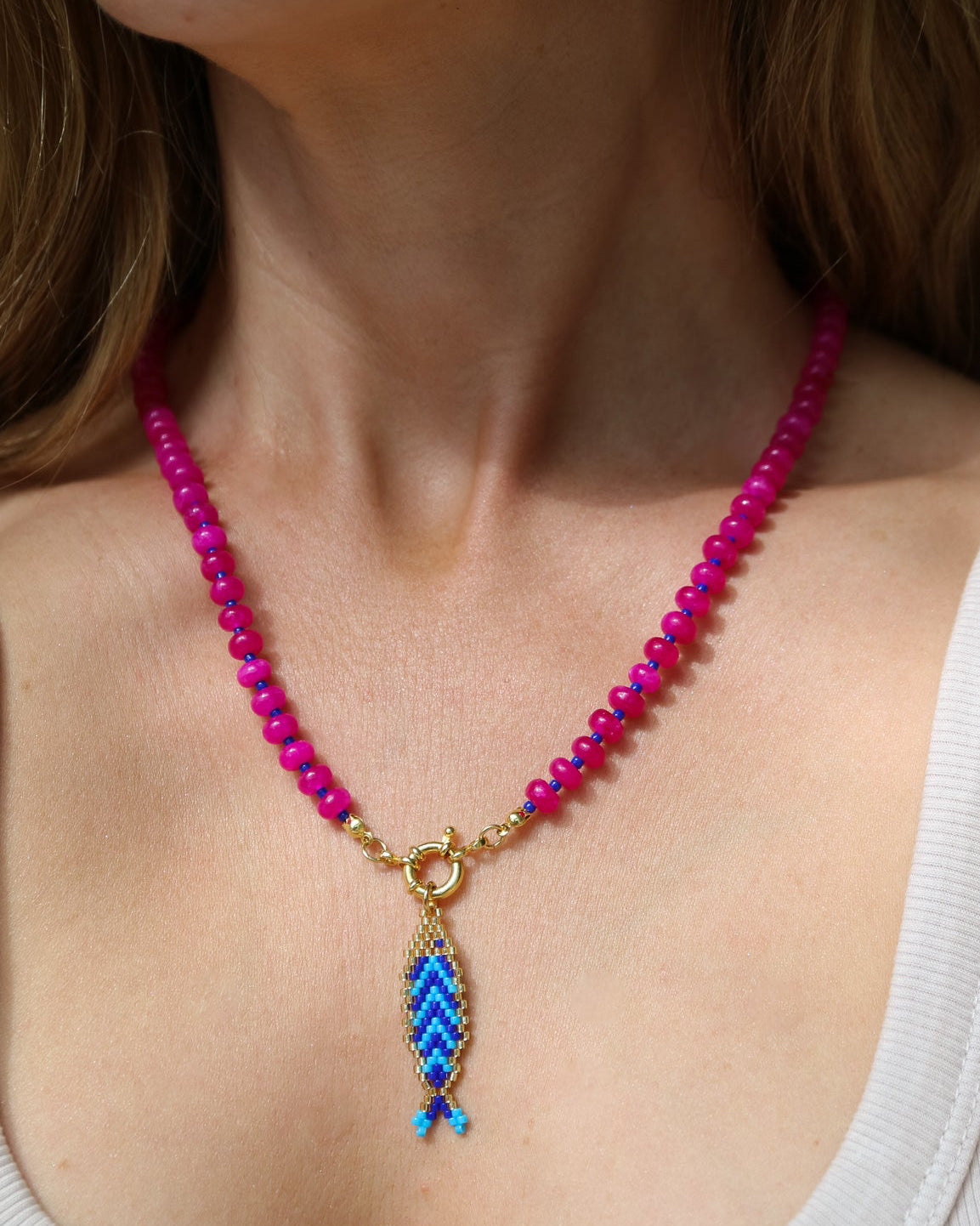 A close-up of a woman wearing a beaded necklace with bright pink beads, a gold clasp, and a blue and purple woven feather-like pendant.