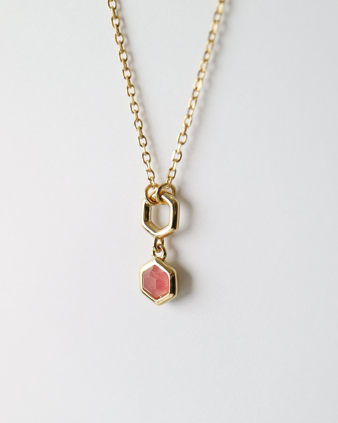 A gold chain necklace featuring a small hexagonal gold pendant with a pink stone inlay, displayed against a clean white background.