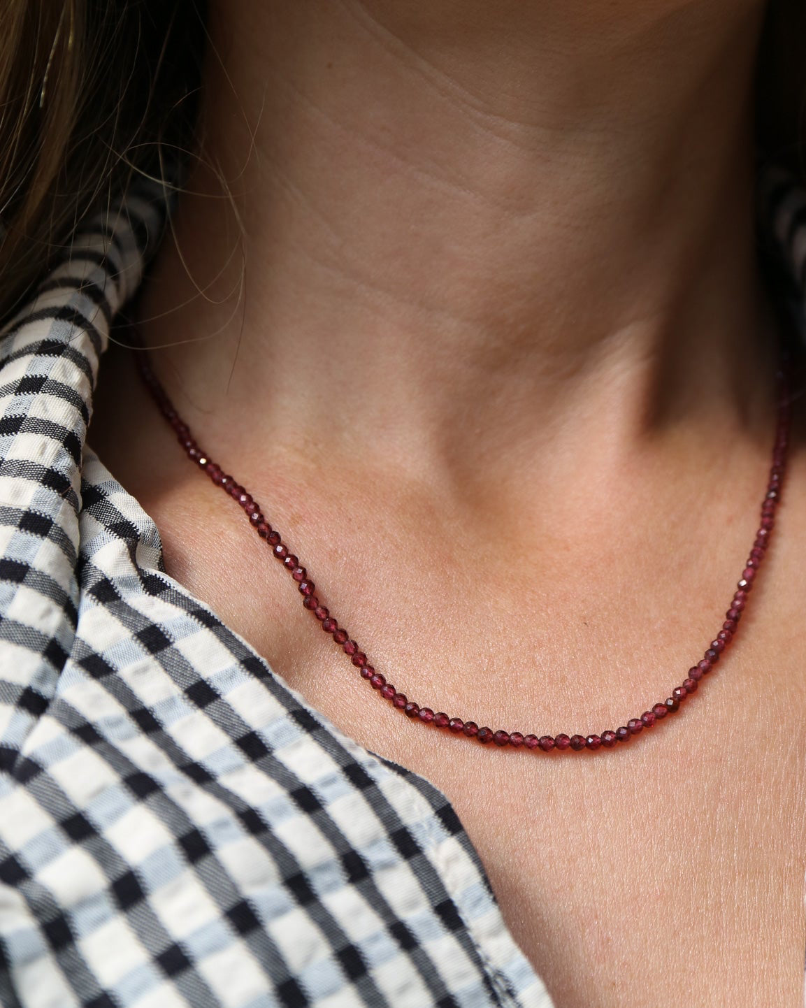 A close-up of a person's neck and chest wearing a delicate dark red beaded necklace over a black and white gingham shirt.