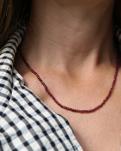 A close-up of a person's neck and chest wearing a delicate dark red beaded necklace over a black and white gingham shirt.