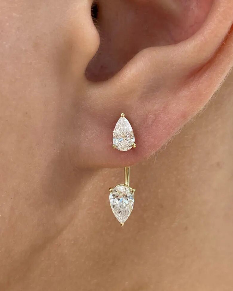 A close-up of a person's ear wearing a pair of gold stud earrings with dangling pear-cut clear gemstones.