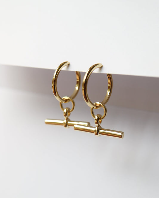 Gold hoop earrings with a small cylindrical charm on a white background
