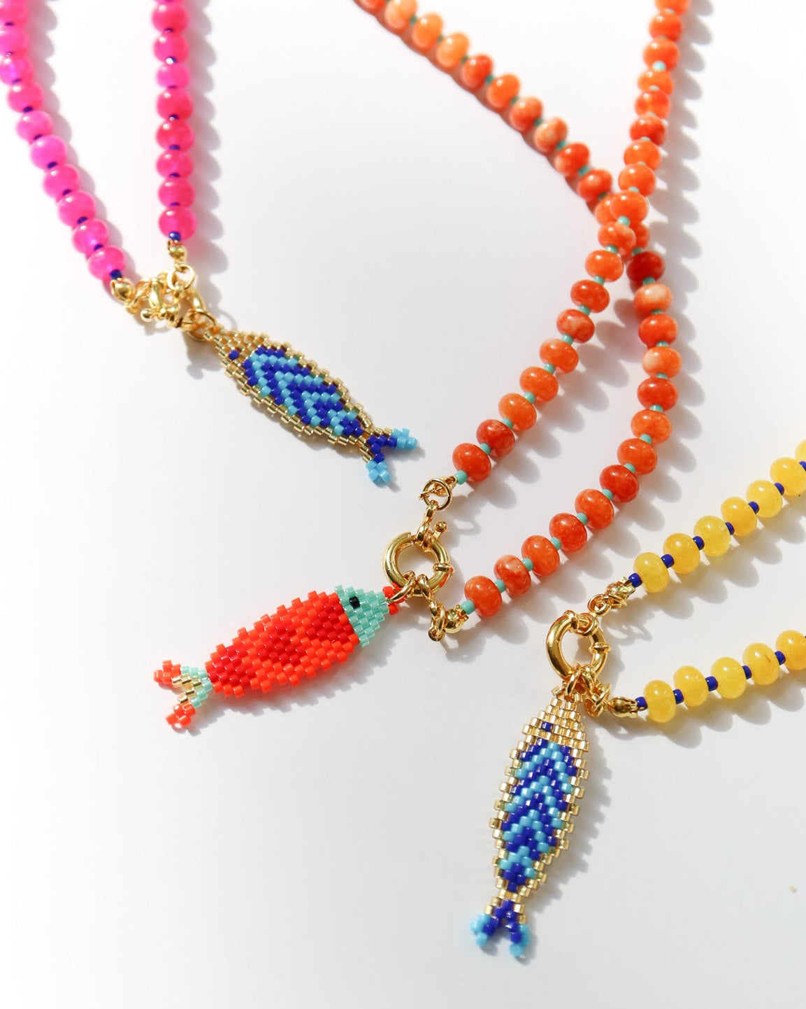 Three beaded necklaces with gold-tone fish charms are arranged on a white background. One necklace is bright pink, one is orange, and one is yellow, each featuring a small woven fish pendant in blue, red, and turquoise.