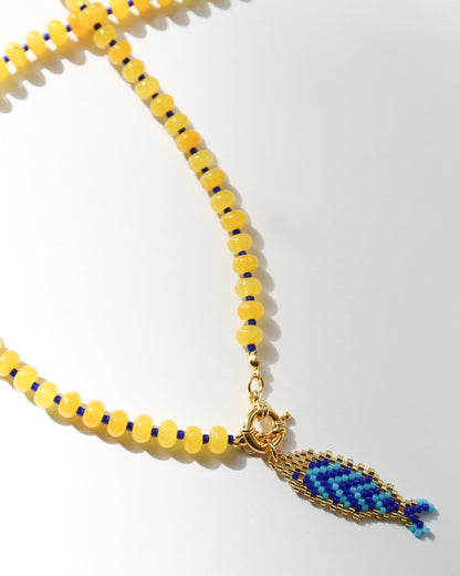 A yellow beaded necklace with a gold-tone clasp and a blue and yellow woven fish-shaped pendant, photographed on a clean white background.