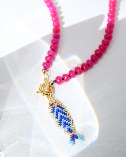 A close-up of a beaded necklace with bright pink faceted beads, a gold-toned clasp, and a dangling fish-shaped charm woven in blue and purple with gold-toned outlines, photographed on a white surface with light reflections.