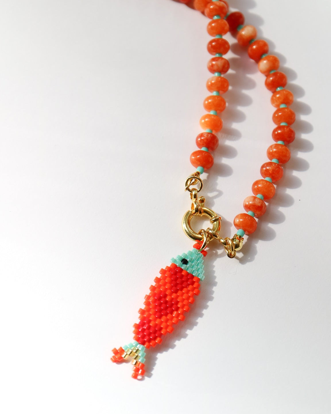 A beaded necklace featuring a strand of orange and red marbled beads with a small fish-shaped charm in red, orange, and light blue, attached with gold hardware, displayed on a clean white background.
