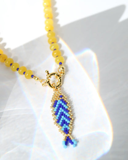 A close-up of a beaded necklace with round yellow beads and a gold-tone fish-shaped pendant filled with blue and lighter blue seed beads, photographed on a white surface with bright light reflections.