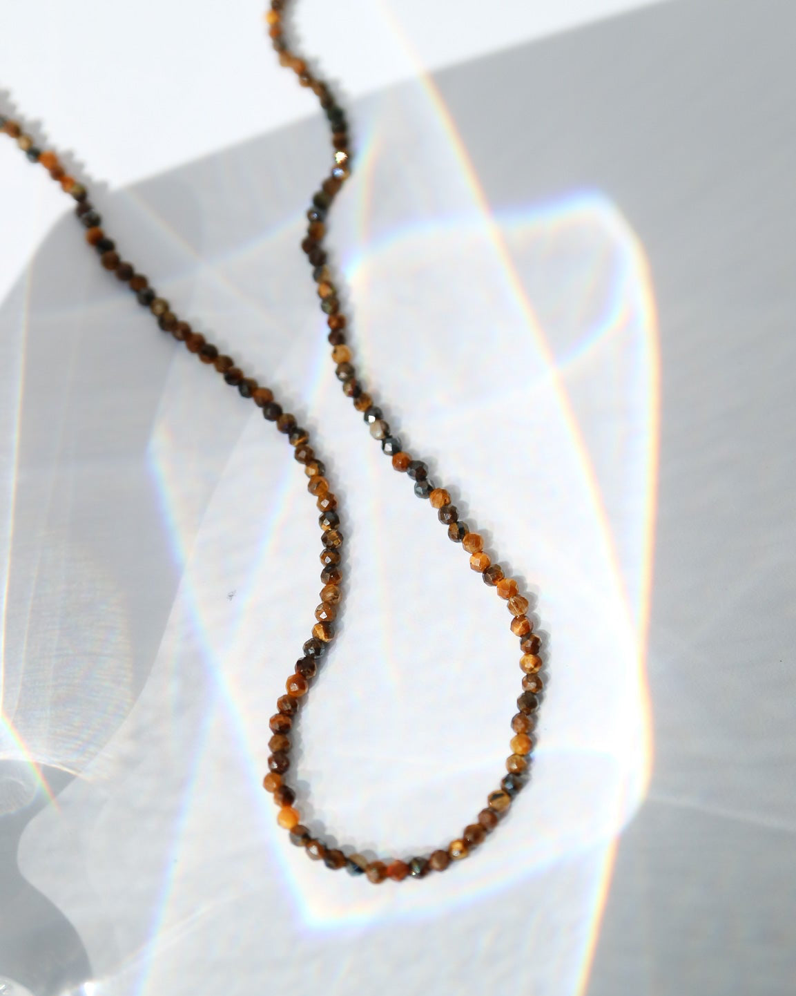 A close-up of a delicate beaded necklace with small brown and amber beads, draped on a white surface with bright light reflections.