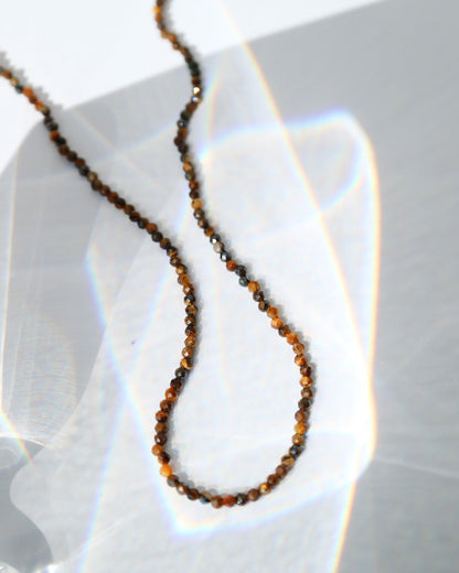 A close-up of a delicate beaded necklace with small brown and amber beads, draped on a white surface with bright light reflections.