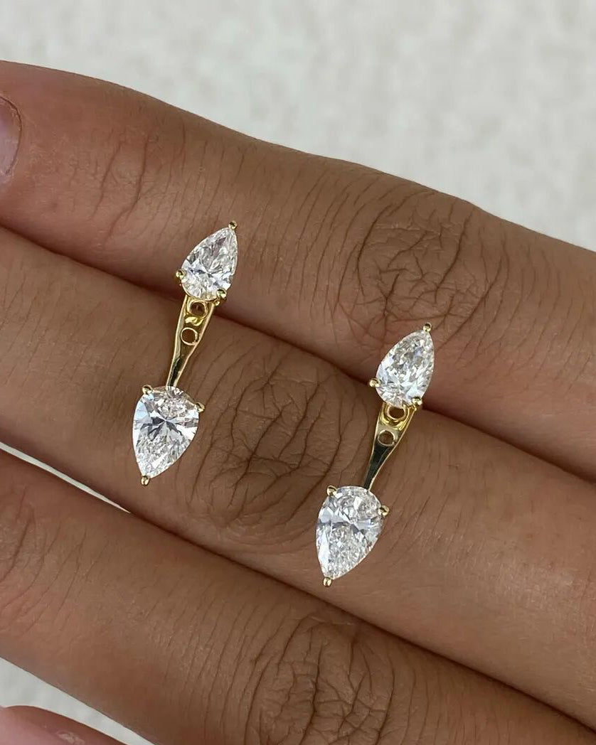 A close-up of a hand wearing a pair of gold earrings with pear-cut clear gemstones, set against a light textured background.