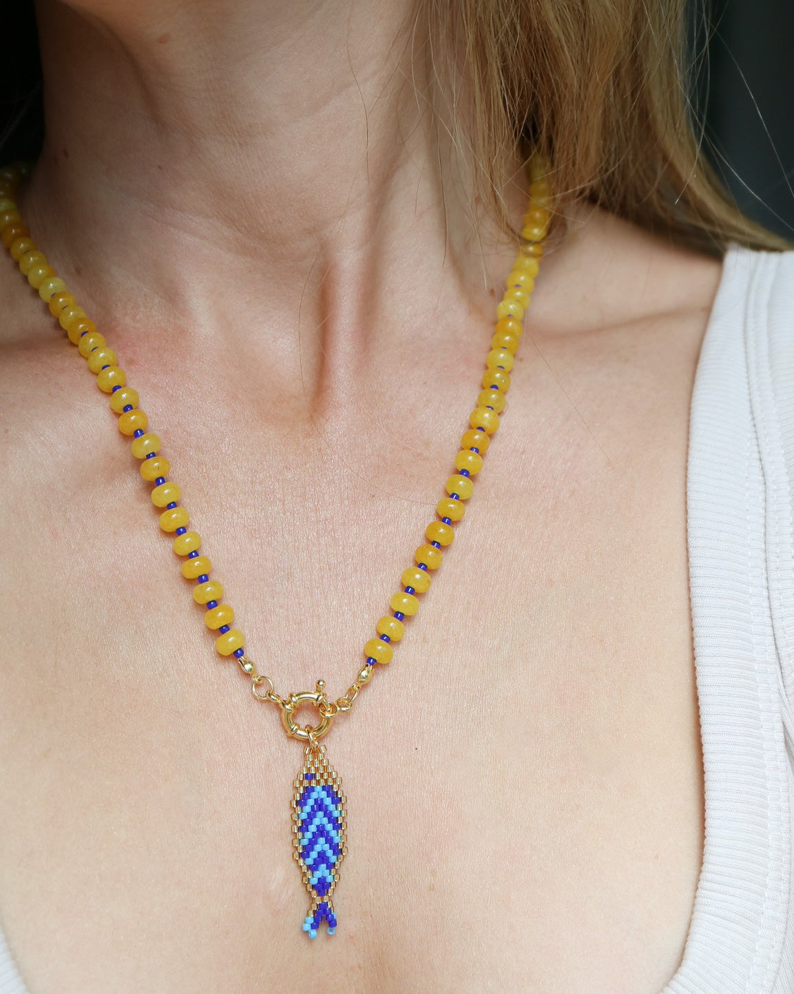 A close-up of a person wearing a gold beaded necklace with a blue and white patterned fish-shaped pendant.