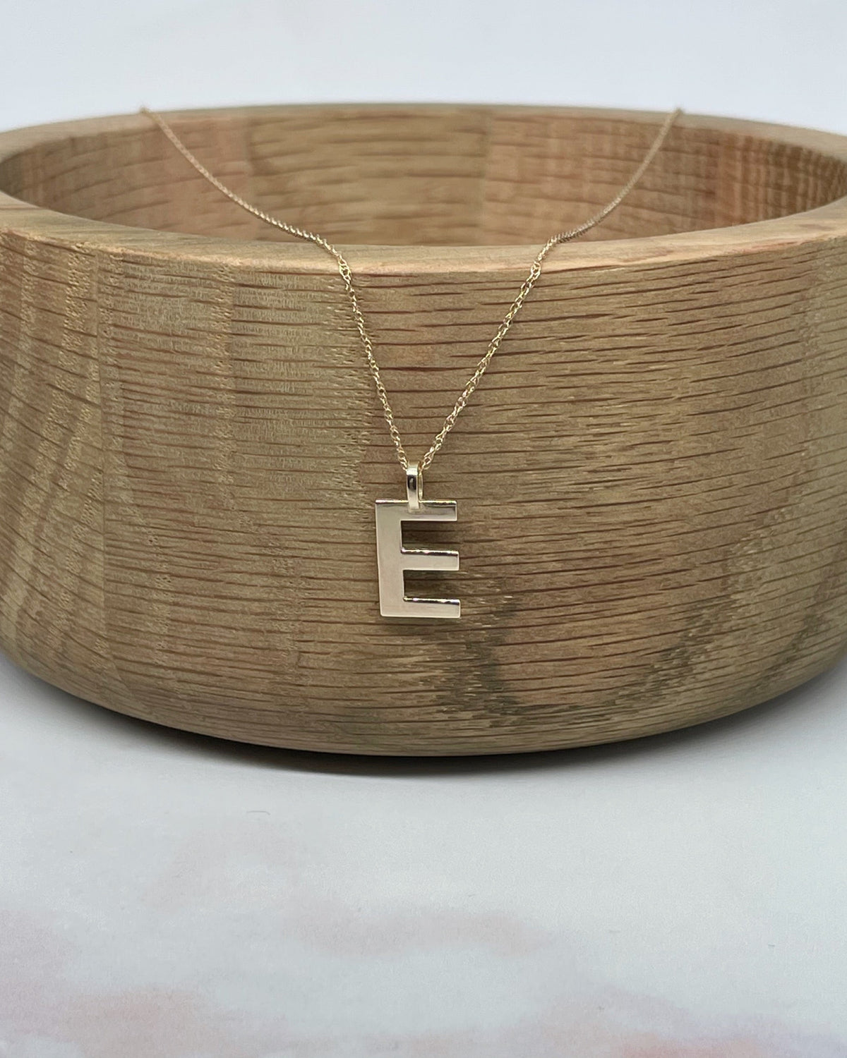 Collective & Co. Solid 9kt E Initial pendant shown on gold chain, draped over wooden bowl